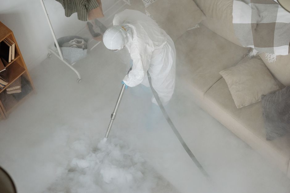 A professional cleaner wearing a black uniform and black gloves is performing surface cleaning on a wooden dining table in a living space. The table appears to be well-maintained and free of dust, wit