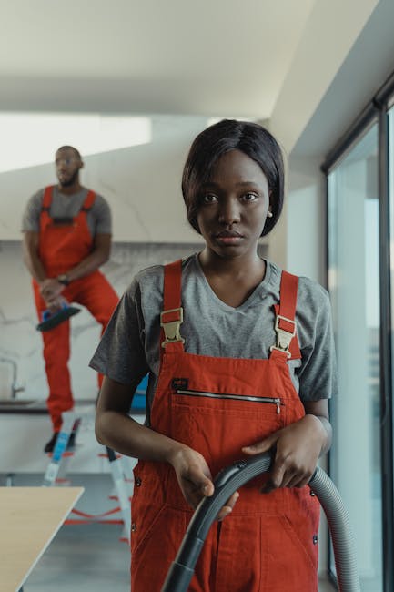 Two professional cleaners in grey shirts and orange overalls are performing surface cleaning in a modern living room with large glass sliding doors. The cleaner on the left is vacuuming the light grey