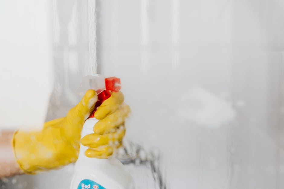 A person wearing bright pink rubber gloves is holding a blue spray bottle of cleaning solution in their right hand while wiping the surface of a white bathroom vanity or cabinet. The surface appears t