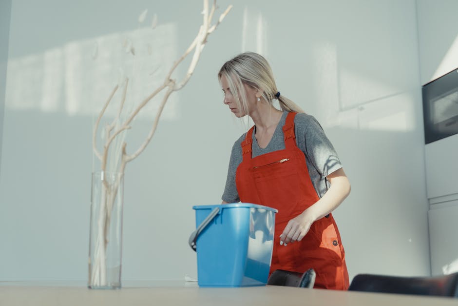 Two professional cleaners, one with dark hair and the other with blonde hair, standing back-to-back against a plain white wall. Both are wearing beige uniforms with short sleeves and appear cheerful. 