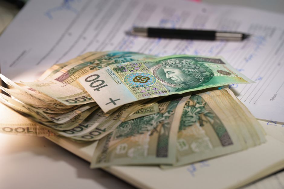 A close-up view of a person’s hand placing a €50 banknote into a black plastic cash box, which contains other euro banknotes and coins scattered around. The background features various financial c